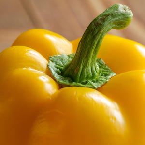 Puede incluir: Un pimiento amarillo brillante con una piel lisa y lustrosa y un tallo verde firme y texturizado en la parte superior. La verdura presenta l&oacute;bulos redondeados y un color vibrante y saturado, mostrando su aspecto fresco y su forma natural.