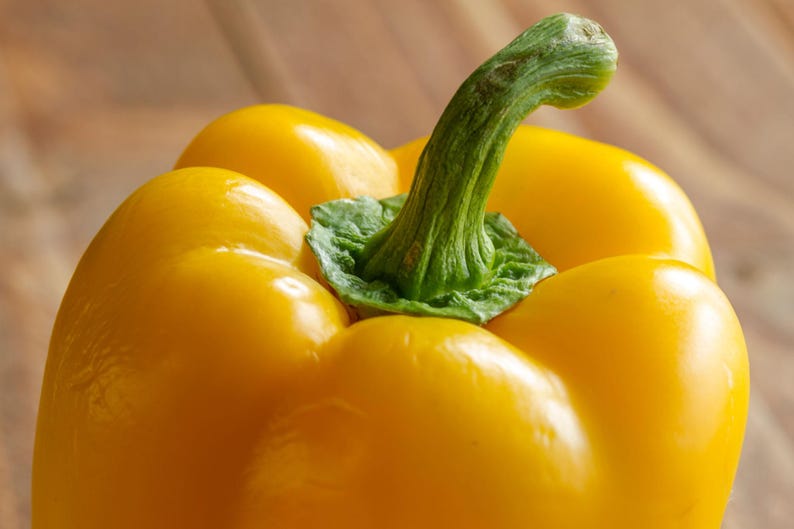 Puede incluir: Un pimiento amarillo brillante con una piel lisa y lustrosa y un tallo verde firme y texturizado en la parte superior. La verdura presenta l&oacute;bulos redondeados y un color vibrante y saturado, mostrando su aspecto fresco y su forma natural.