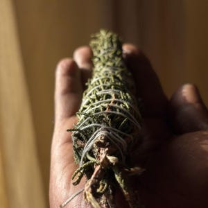 May include: A bundle of dried green cedar foliage tightly wrapped with thin white cotton string. The cylindrical arrangement features textured, needle-like leaves and woody stems, secured in a spiral pattern along the length of the botanical bundle.