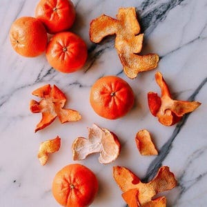 May include: Five bright orange tangerines arranged alongside several pieces of dried, textured orange peel. The fruit is round and smooth, while the peels show a rough, porous interior surface and a vibrant, slightly wrinkled exterior texture.
