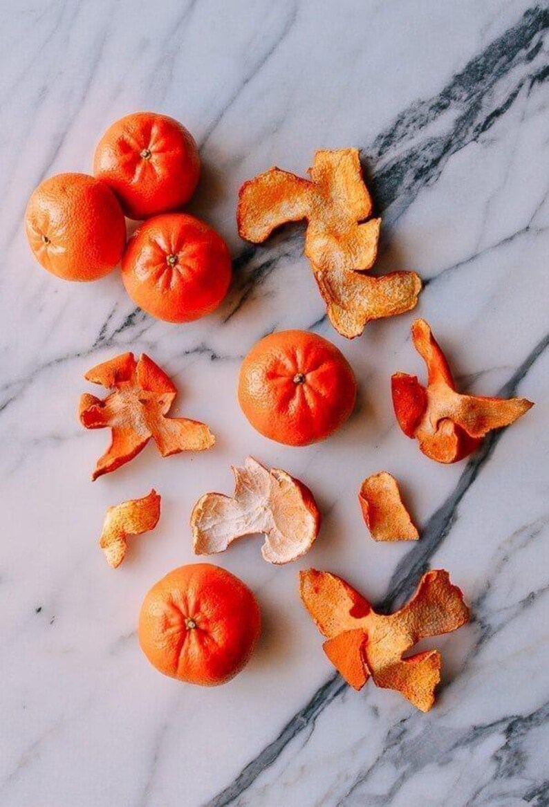May include: Five bright orange tangerines arranged alongside several pieces of dried, textured orange peel. The fruit is round and smooth, while the peels show a rough, porous interior surface and a vibrant, slightly wrinkled exterior texture.