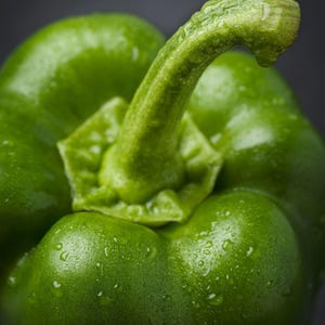 Puede incluir: Un pimiento verde vibrante con una piel lisa y brillante cubierta de peque&ntilde;as gotas de agua. La verdura tiene un tallo curvo prominente que se extiende desde el centro superior, con una estructura firme y lobulada de color verde intenso.