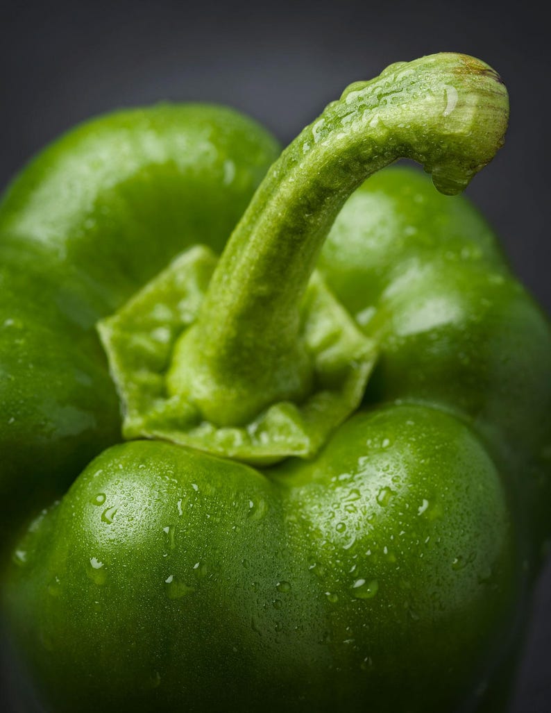 Puede incluir: Un pimiento verde vibrante con una piel lisa y brillante cubierta de peque&ntilde;as gotas de agua. La verdura tiene un tallo curvo prominente que se extiende desde el centro superior, con una estructura firme y lobulada de color verde intenso.