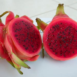 May include: A dragon fruit cut in half, revealing a vibrant magenta interior speckled with tiny black seeds. The outer skin is a bright pinkish-red with green, leaf-like scales. The fruit is presented on a white surface with thin black lines.