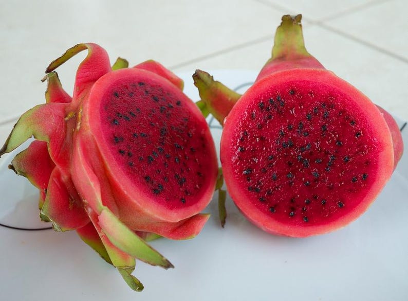 May include: A dragon fruit cut in half, revealing a vibrant magenta interior speckled with tiny black seeds. The outer skin is a bright pinkish-red with green, leaf-like scales. The fruit is presented on a white surface with thin black lines.