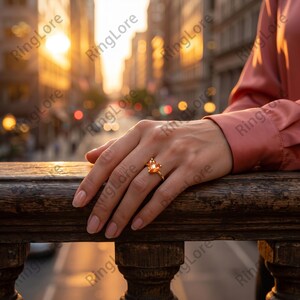 May include: A gold ring with a central orange gemstone and smaller surrounding stones. The ring is on a person's finger, resting on a wooden railing. The background features a blurred cityscape with warm, golden light.