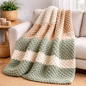 May include: A cozy, handmade crochet blanket draped over a beige sofa. The blanket features horizontal stripes of cream, tan, and sage green. The textured stitches create a soft, inviting appearance, perfect for home decor and comfort.