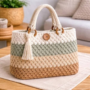 May include: A handmade crochet tote bag with a striped pattern in cream, green, tan, and brown. The bag features wooden handles, a wooden button closure, and a cream-colored tassel. The bag is sitting on a wooden table.