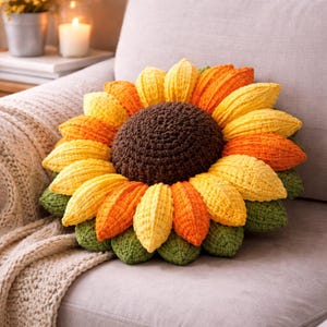 May include: A crocheted sunflower pillow with a brown center and yellow, orange, and green petals. The pillow is placed on a gray couch with a cream-colored knitted throw blanket.