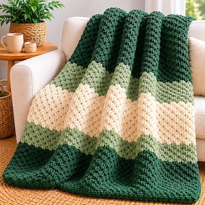 Puede incluir: Una manta de ganchillo gruesa con rayas horizontales en verde bosque oscuro, verde salvia y crema. La manta presenta un patrón de puntos en relieve por toda la superficie y está colocada sobre el brazo de un sofá tapizado de color claro.