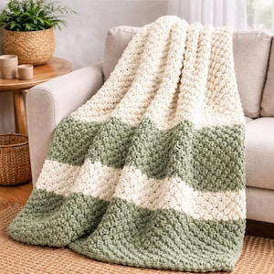 Könnte beinhalten: Eine grob gestrickte Wohndecke mit abwechselnden horizontalen Streifen aus cremefarbenem und salbeigrünem Garn. Die Decke weist ein durchgehendes, dickes und strukturiertes Noppenmuster auf, das ihr ein weiches und voluminöses Aussehen verleiht.