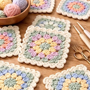 May include: A collection of colorful, handmade crochet coasters in pastel shades of pink, yellow, blue, and green. Each square coaster features a floral design and a scalloped edge. Crochet hooks and yarn balls are also visible.