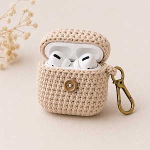 Może przedstawiać: Beżowe, szydełkowe etui na AirPods z drewnianym zapięciem na guzik i złotym karabińczykiem. Etui jest otwarte, ukazując białe słuchawki AirPods. Etui ma chronić i przenosić bezprzewodowe słuchawki.
