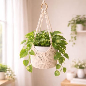 Może przedstawiać: Kremowy makramowy uchwyt na rośliny, zawieszony na drewnianym pierścieniu, trzymający białą doniczkę z zieloną rośliną liściastą. Tkany bawełniany uchwyt na rośliny jest przeznaczony do użytku w pomieszczeniach.