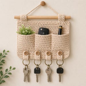 May include: A beige crochet key holder with four pockets and key rings. The key holder has a wooden dowel and a wooden button closure. The pockets contain a small plant, a car key, and a pen. Keys hang from the bottom.