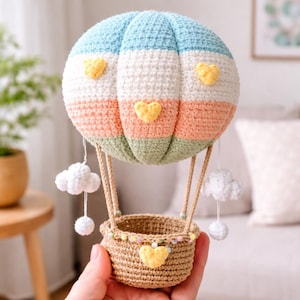 Può includere: Una mongolfiera all'uncinetto fatta a mano in colori pastello. Il pallone presenta strisce blu, bianche, pesca e verdi, con accenti a cuore giallo. Un cestino marrone è sospeso sotto, con due ornamenti a nuvola bianca appesi.