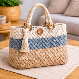 May include: A cream, blue, and tan crochet tote bag with wooden handles and a button closure. The bag features a cream tassel and wooden rings. The bag is made with a textured stitch. The bag is sitting on a neutral-colored surface.
