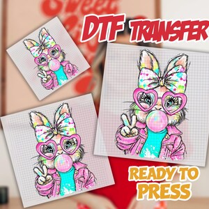 May include: DTF transfer featuring a cartoon bunny with a bow, heart-shaped glasses, and a bubblegum bubble. The bunny is wearing a pink jacket and a teal shirt, giving a peace sign. The words "DTF TRANSFER" and "READY TO PRESS" are visible.
