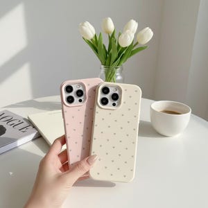May include: Two phone cases, one light pink and one cream, are held in a hand. Both cases have a pattern of small gray hearts. The cases are next to a vase of white tulips and a cup of coffee.