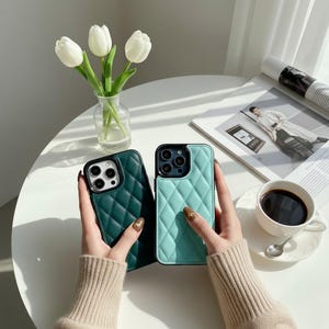 May include: Two quilted phone cases, one teal and one dark green, are held up. The cases have a diamond pattern and black borders. A vase of white tulips, a cup of coffee, and a magazine are also visible.