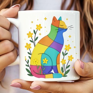 May include: White ceramic mug featuring a colorful patchwork cat design. The cat is composed of various colored squares and is surrounded by yellow flowers and stars. The mug has a curved handle and is held by a person.