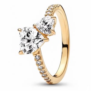May include: A gold-colored ring featuring two heart-shaped, clear gemstones. The band is also adorned with smaller, clear gemstones. The ring is set against a white background, highlighting its design and sparkle.
