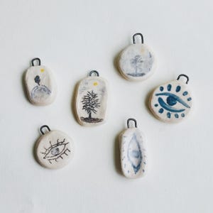 May include: Six off-white ceramic pendants with dark metal loops. The pendants feature hand-painted designs including trees, suns, and stylised eyes in blue, black, and yellow tones. Each piece has an irregular, organic shape and a smooth finish.