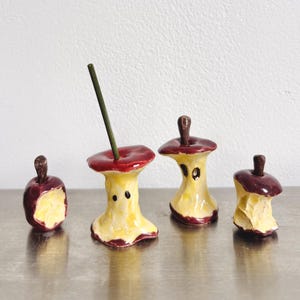 May include: Four ceramic apple core sculptures with glossy red skin and yellow flesh. Each piece features a brown stem and irregular, bitten edges. One core includes a thin green incense stick inserted into the top centre of the fruit.