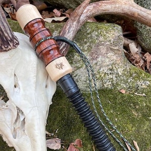 May include: A hunting call with a wooden barrel and a black, ribbed bellow. The barrel features alternating light and dark wood rings. A green and black lanyard is attached. The call rests on a mossy rock near a deer skull and antlers.