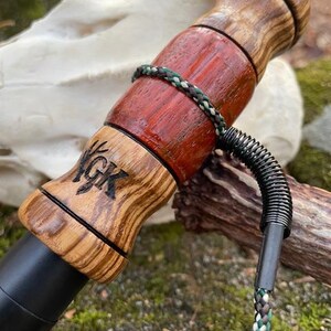 May include: A wooden hunting call with a black mouthpiece and a decorative wooden body. The body features a reddish-brown center section and a lighter wood grain pattern on the ends. A braided cord and a metal coil are attached. The letters "YGK" are visible.