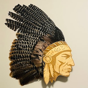 Puede incluir: Perfil de madera de un nativo americano con un tocado de plumas. El perfil es de color marrón claro con detalles grabados, incluyendo un tocado y plumas. El tocado está hecho de plumas marrones oscuras y negras.