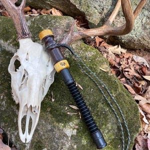 May include: A hunting call with a black, ribbed body and wooden accents. The call is dark brown and tan, with a lanyard. The call is resting on a mossy rock next to a deer skull and antlers.