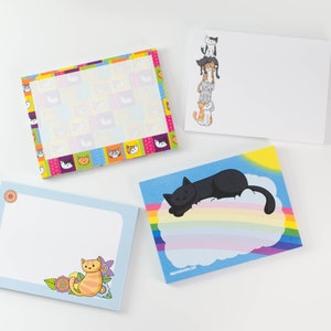 May include: Four different cat-themed notepads. One notepad has a rainbow and a black cat sleeping on a cloud. Another notepad has a colorful patchwork design with cat faces. A third notepad has a blue background with a single orange cat and flowers. The final notepad has a white background with a stack of five cats.