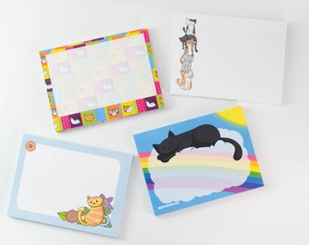May include: Four different cat-themed notepads. One notepad has a rainbow and a black cat sleeping on a cloud. Another notepad has a colorful patchwork design with cat faces. A third notepad has a blue background with a single orange cat and flowers. The final notepad has a white background with a stack of five cats.