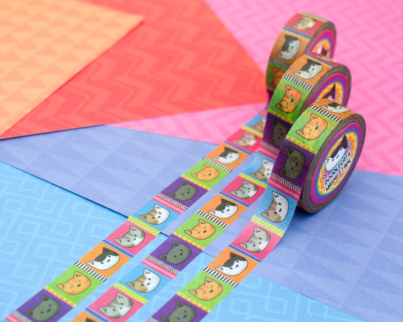 May include: Colorful washi tape rolls with a repeating pattern of cartoon cats' faces. The tape is unrolled on a blue background and shows the pattern of the tape.