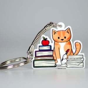May include: Orange and white cat keychain with a red pen and a stack of books. The cat is sitting on a stack of papers and holding a red pen. The keychain has a silver spring ring.