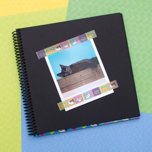 May include: A black spiral bound scrapbook with a photo of a black cat lying on a wooden surface. The photo is decorated with colorful cat face stickers.