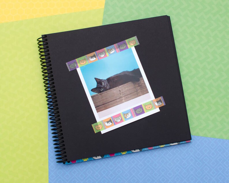 May include: A black spiral bound scrapbook with a photo of a black cat lying on a wooden surface. The photo is decorated with colorful cat face stickers.