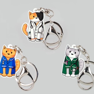 May include: Three cat keychains with different coloured scrubs and a stethoscope. The cats are orange and white, orange and blue, and grey and green.