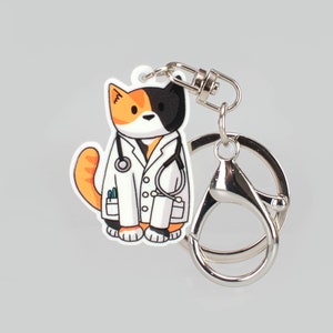 May include: A black, white and orange cat wearing a white doctor's coat with a stethoscope around its neck. The cat is attached to a silver key chain.