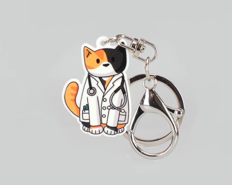 May include: A black, white and orange cat wearing a white doctor's coat with a stethoscope around its neck. The cat is attached to a silver key chain.