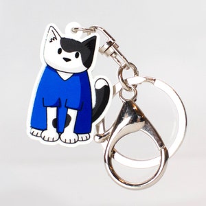 May include: A black and white cat wearing a blue jumpsuit keychain. The keychain is attached to a silver metal ring.