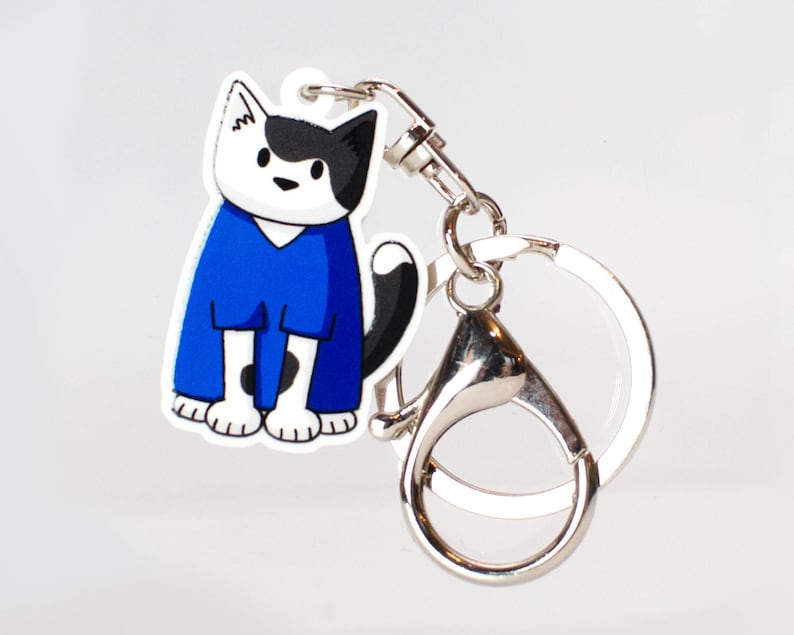 May include: A black and white cat wearing a blue jumpsuit keychain. The keychain is attached to a silver metal ring.