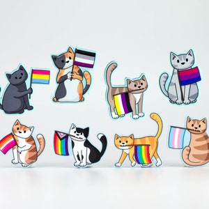 May include: Set of nine colourful cat stickers with pride flags. Each cat is holding a different flag, including the bisexual, transgender, lesbian, pansexual, and genderfluid flags.