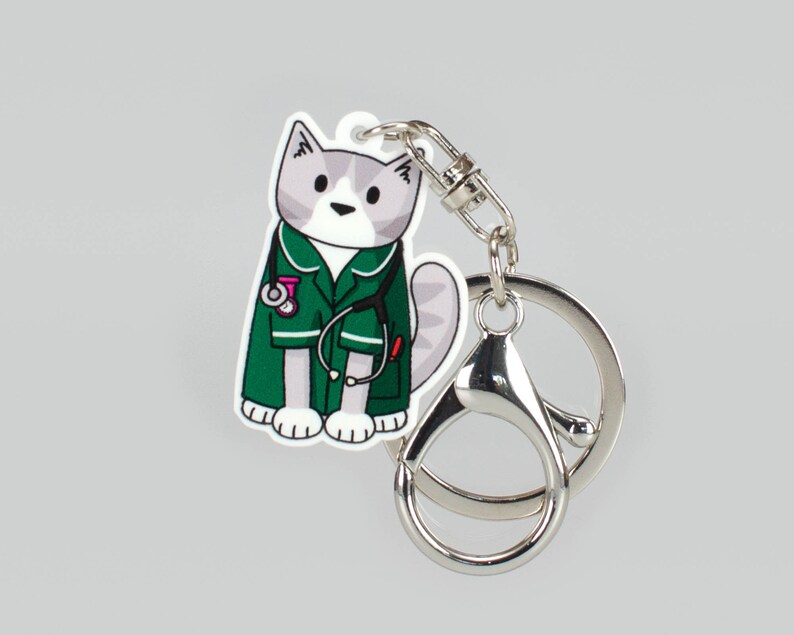 May include: A white and grey cat wearing a green medical scrubs uniform with a stethoscope and a pink bandage. The cat is holding a small white medical syringe. The cat is attached to a silver key chain.