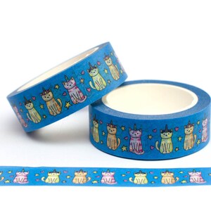 May include: Two rolls of blue washi tape with a repeating pattern of colorful cats wearing unicorn horns. The tape is decorated with stars and hearts. A third roll of tape is unrolled on a white surface, showing the pattern.