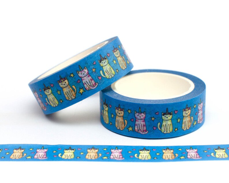 May include: Two rolls of blue washi tape with a repeating pattern of colorful cats wearing unicorn horns. The tape is decorated with stars and hearts. A third roll of tape is unrolled on a white surface, showing the pattern.