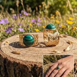 Peut inclure: Deux bagues en or avec des pierres de malachite. L'une a une bande avec des découpes de feuilles, l'autre a une bande avec une pierre de malachite. Les bagues sont présentées sur une surface en bois avec un fond naturel de fleurs et de verdure.