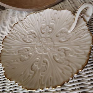 May include: Cream-colored, leaf-shaped serving dish with an ornate floral and scroll design. The dish has a gold-colored trim and a curved handle. A tag with the text "Elmore General Store" is attached to the handle.