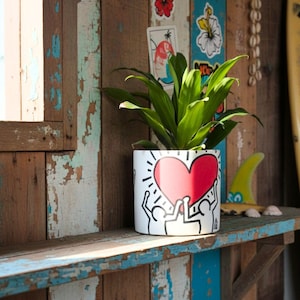 Op de afbeelding: Een witte keramische plantenpot met een rood hartontwerp en zwarte lijntekening, met een groene plant. De pot staat op een verweerde houten plank met blauwe rand, tegen een achtergrond van houten planken en surf-thema stickers.